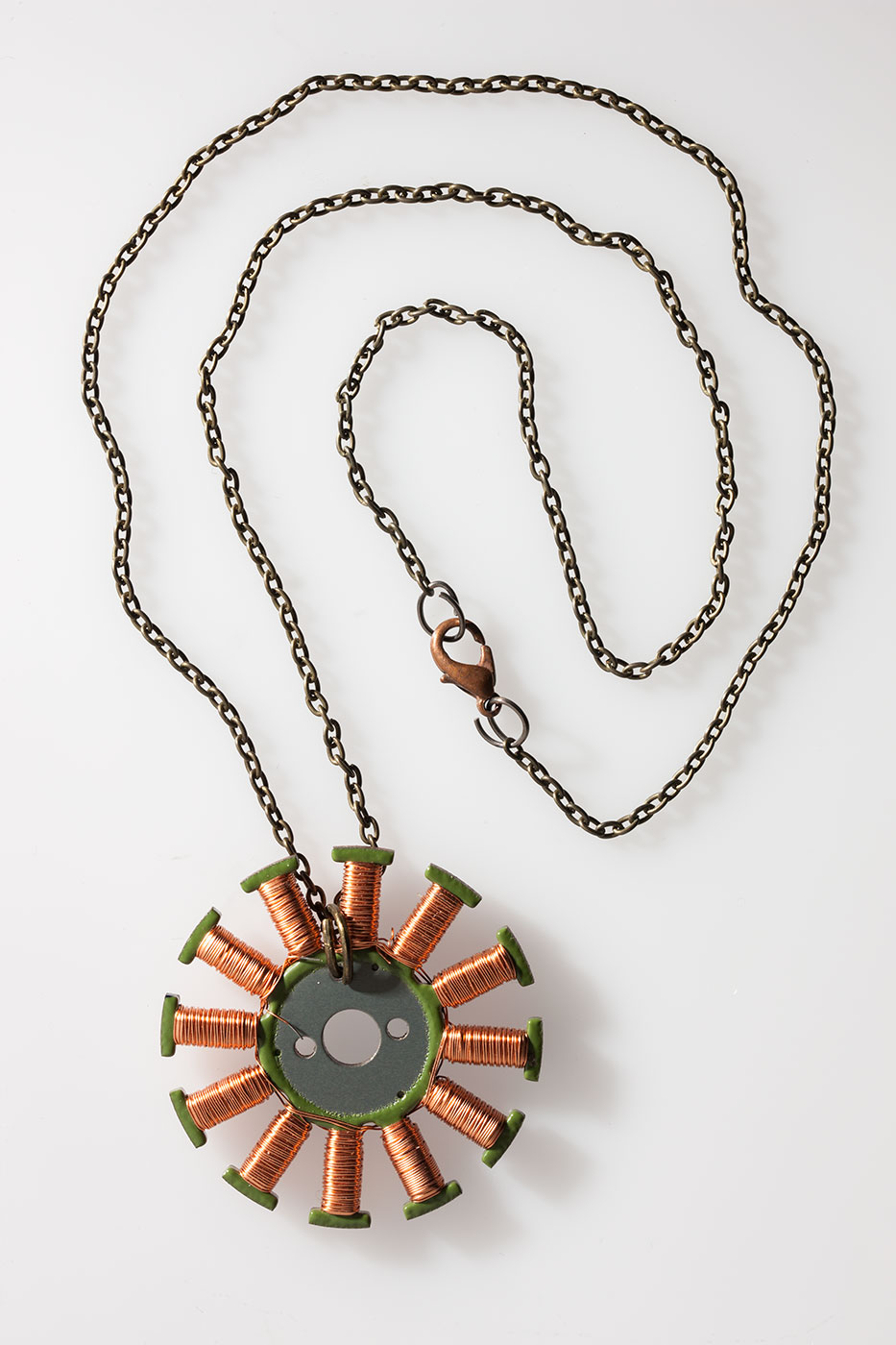 A circular metal pendant with green coloured t-shapes coiled with copper wire radiating from the centre, on a metal chain. - click to view larger image