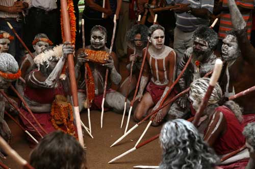 A colour photograph of eleven Australian Indigenous people sitting in a circle. Each of them has traditional body and face markings. Some have white pigment in their hair. Many hold long spears that are painted red and white. The tips of the spears rest on the floor. Behind the group stand other people, only visible from the waist down. - click to view larger image