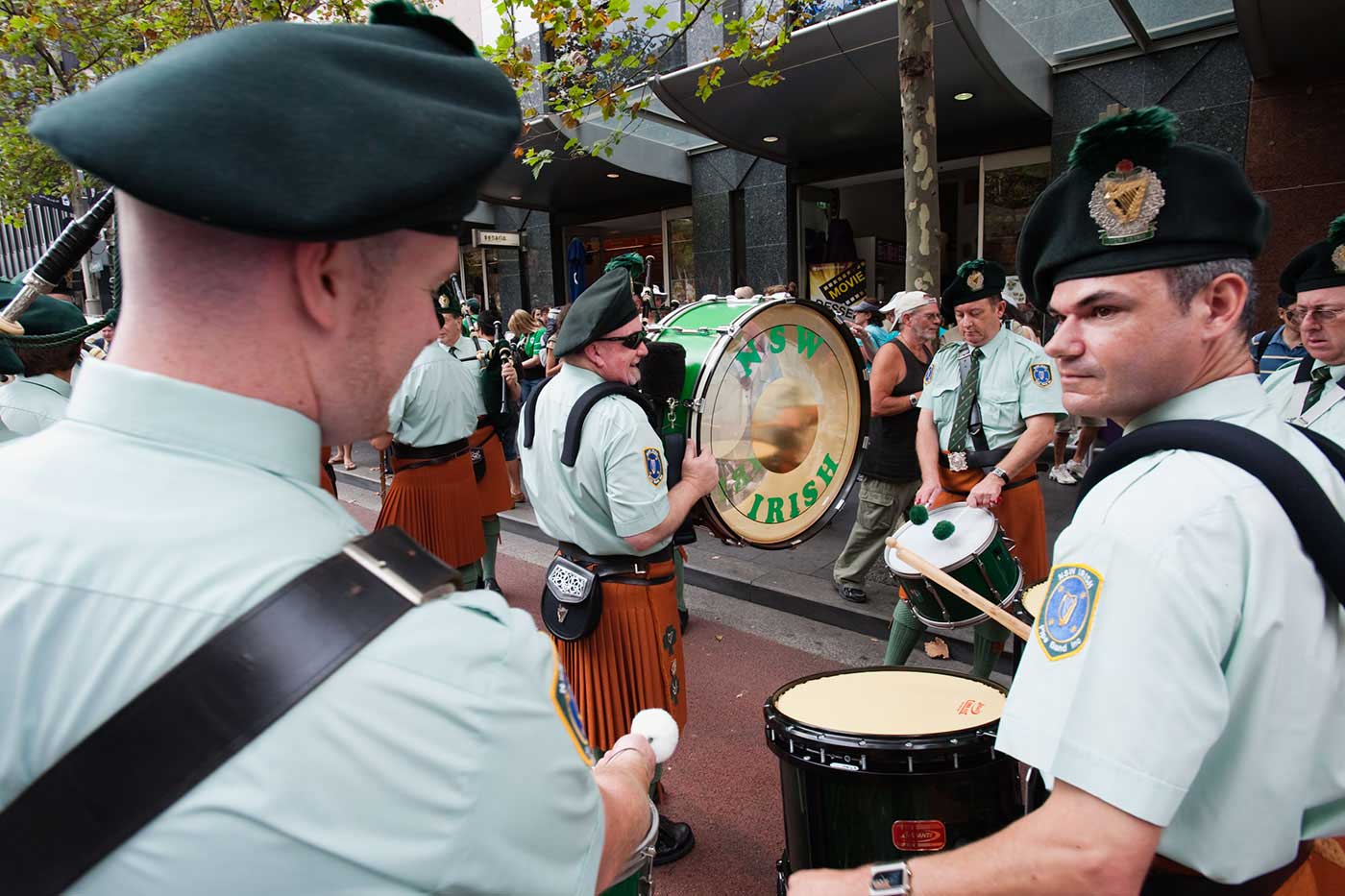 Drumming band of men wearing kilts, shirts and hats in a procession on a city street. - click to view larger image