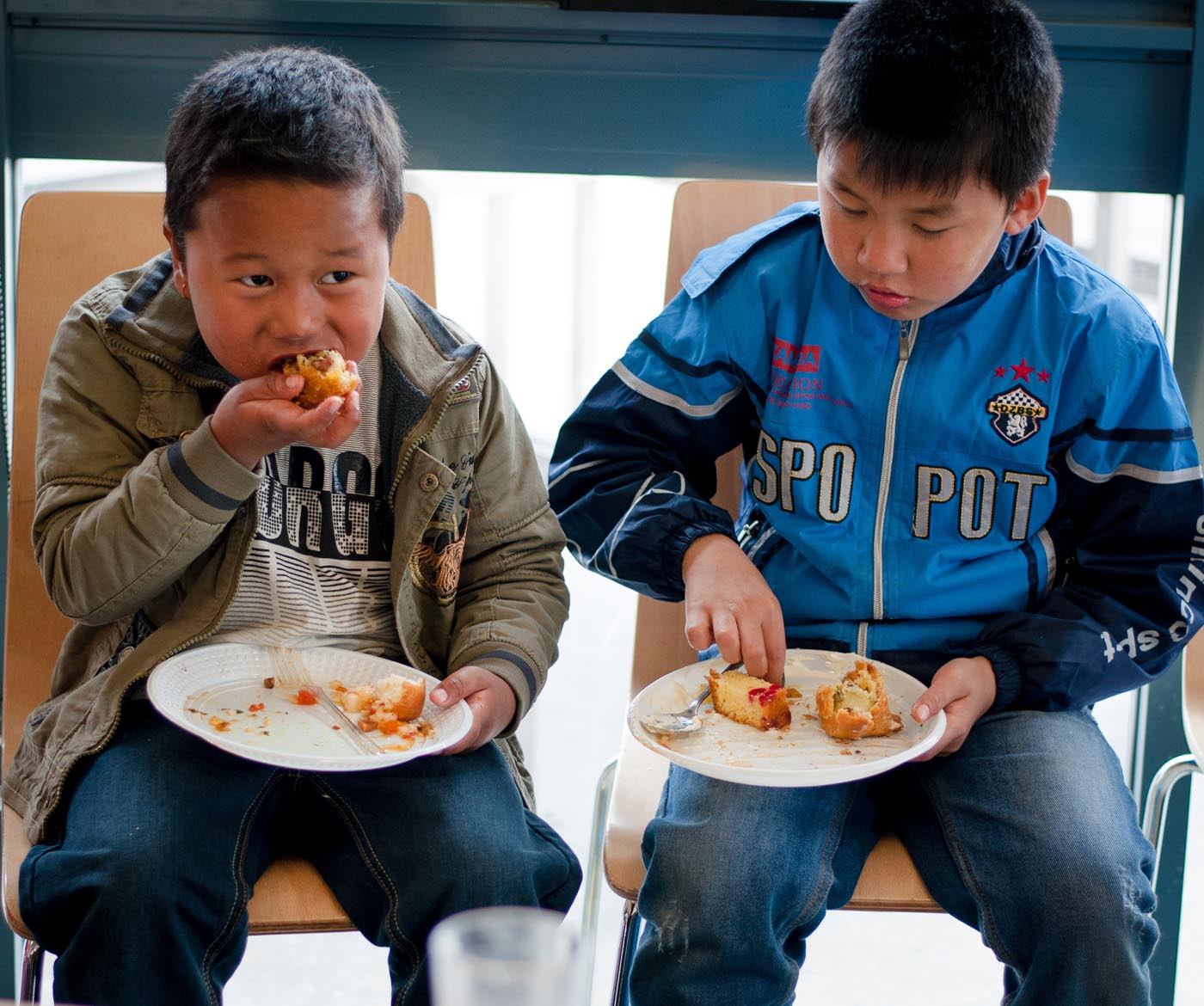 Photo of two young boys sitting down holding plates with various items on them with the boy on the left in the midst of placing an item in his mouth and the boy on the right using his fingers to pick up an item from the plate - click to view larger image