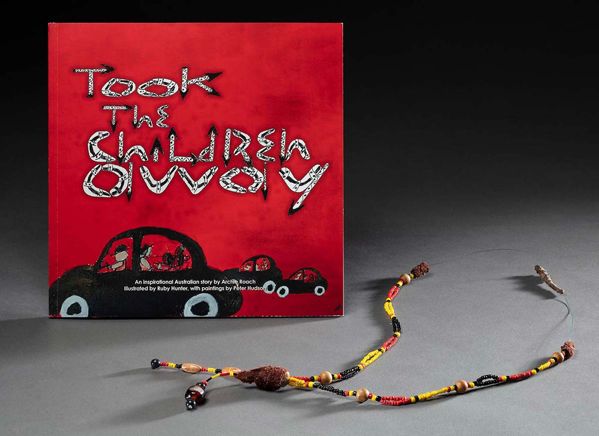 A red, yellow and black necklace with beads, in front of a book with a red cover. The book has an illustration of three black cars with adults in the front seat, children in the back.