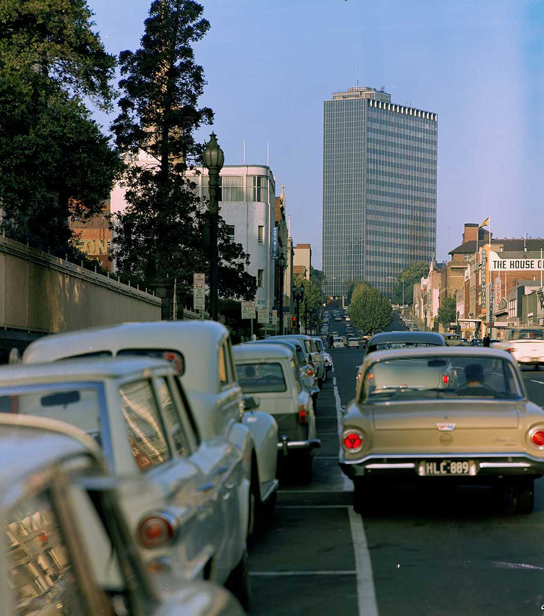 Photo depicting cars from the 1960s on a city road. There is a view of a tall building in the distance. - click to view larger image