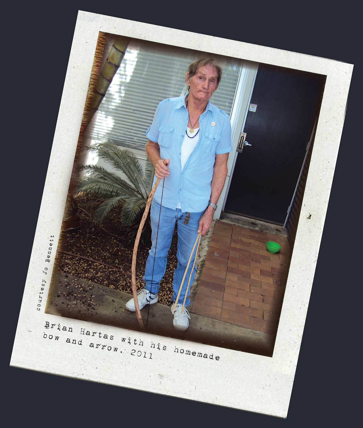 Polaroid photograph showing a man standing and holding a wooden bow in his left hand and two arrows in his right. Typewritten text below reads 'Bryan Hartas with his homemade bow and arrow, 2011'. 'Courtesy Jo Bennett' is written along the left side. - click to view larger image