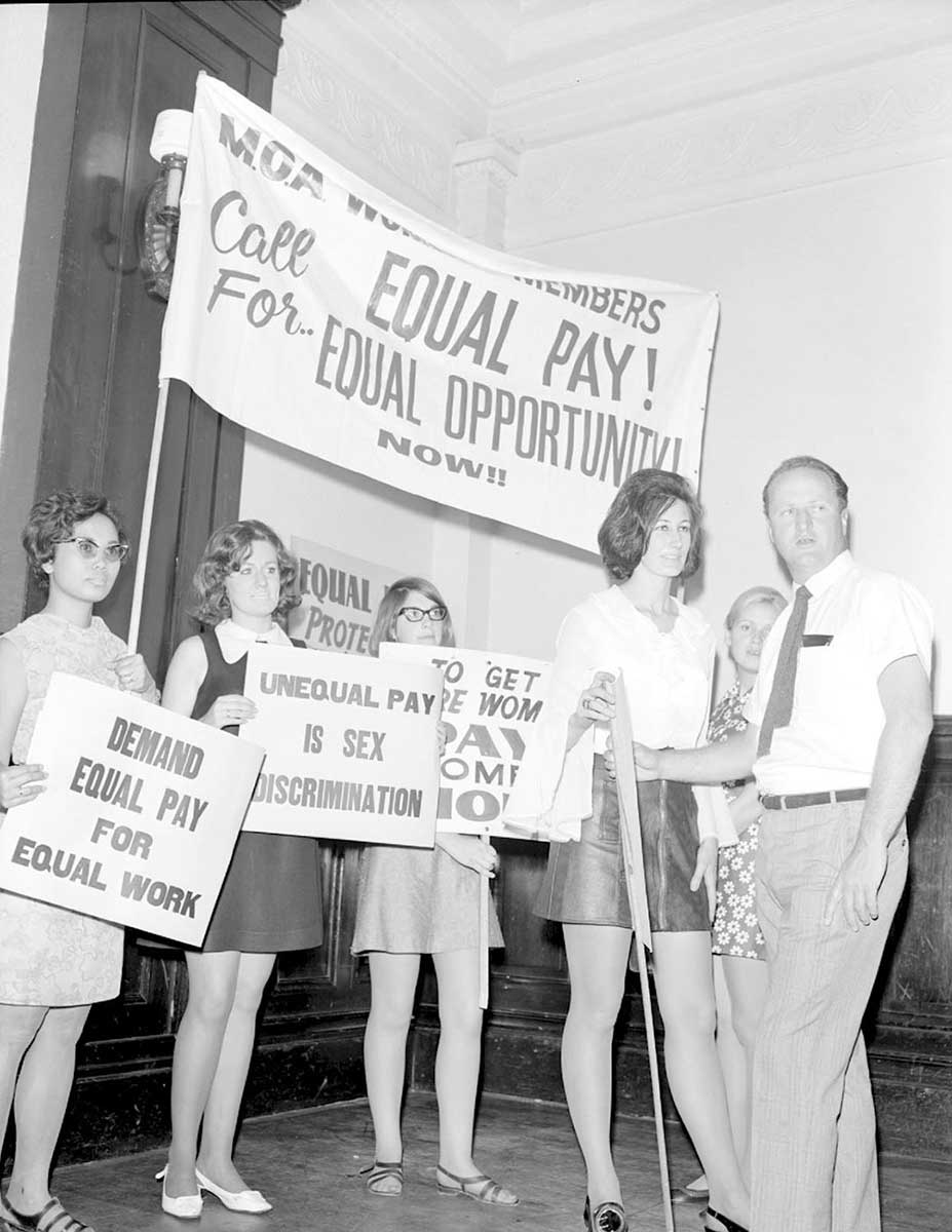 Black-and-white photo of female protesters and one male protester holding up banners. A large banner behind them reads ‘call For EQUAL PAY? EQUAL OPPORTUNITY. Now!!’. - click to view larger image