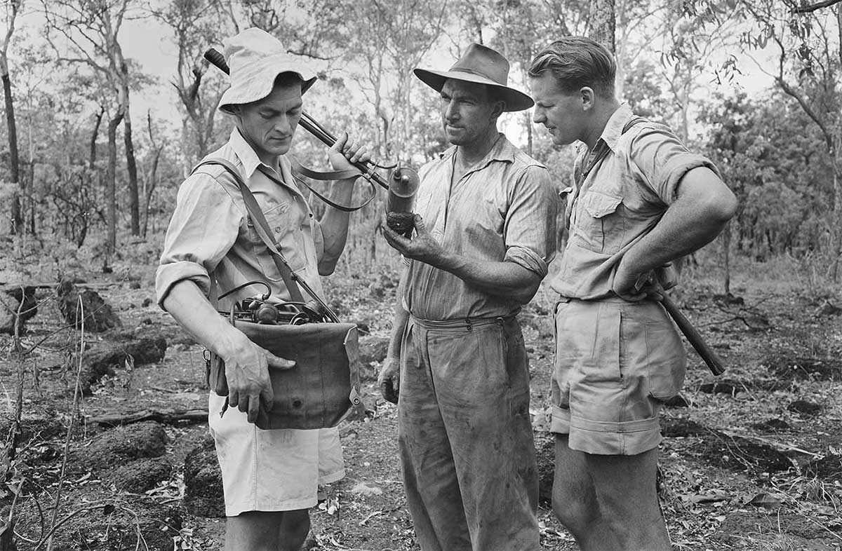 Black and white photo of three men standing in bush land. the man on the left is carrying a Geiger counter and other equipment, the man in the middle is holding a rock or mineral sample in his hand and the man on the right is holding a geological tool.