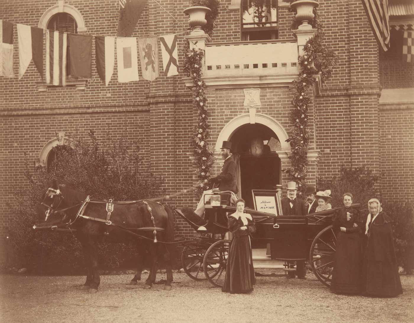 Sepia coloured photograph showing a landau carriage pulled by a horse standing in front of the homestead entrance with the honeymoon couple being greeted by relatives. - click to view larger image