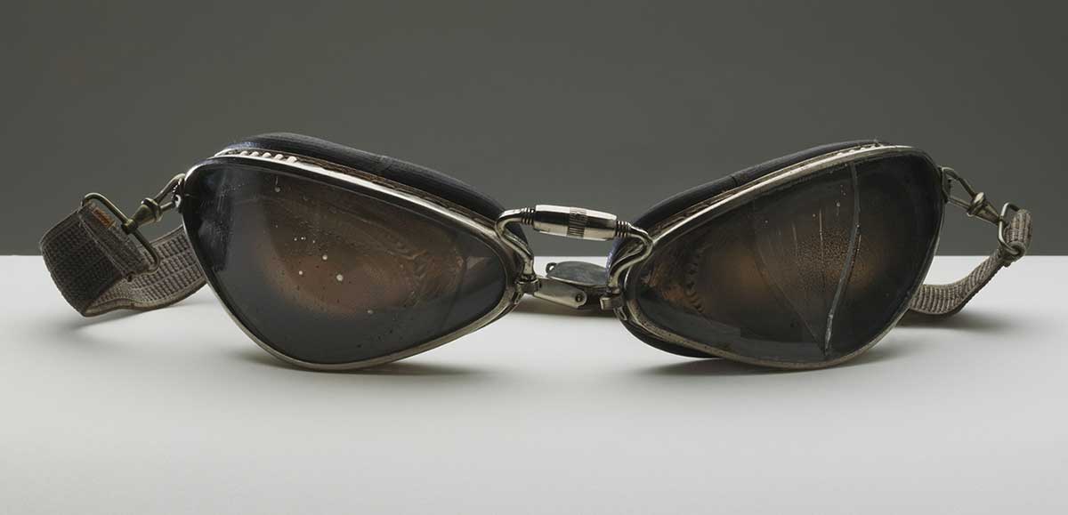Pair of driving goggles owned by Joan Richmond during her racing career. The goggles have metal rimmed eyepieces, each with rubber seals attached to the back, an elastic strap at the back with metal adjustment clips, and a metal piece in the front centre which allows for adjustment. The left eyepiece is damaged with a section missing.