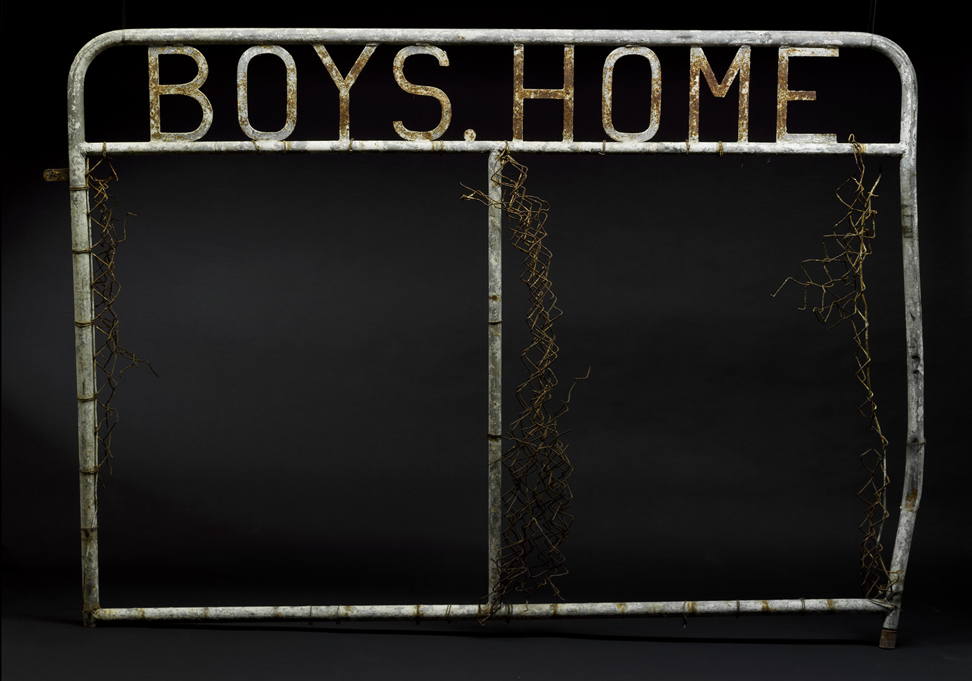 A metal gate panel with welded metal letters at the top which read 'BOYS HOME'. Remnants of wire mesh are attached to the tubular upright components of the gate, which are on the outer edges and in the centre. - click to view larger image