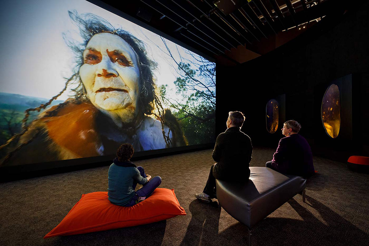 Three museum visitors are seated in front of a large digital screen featuring a person wearing animal skins and a painted face.