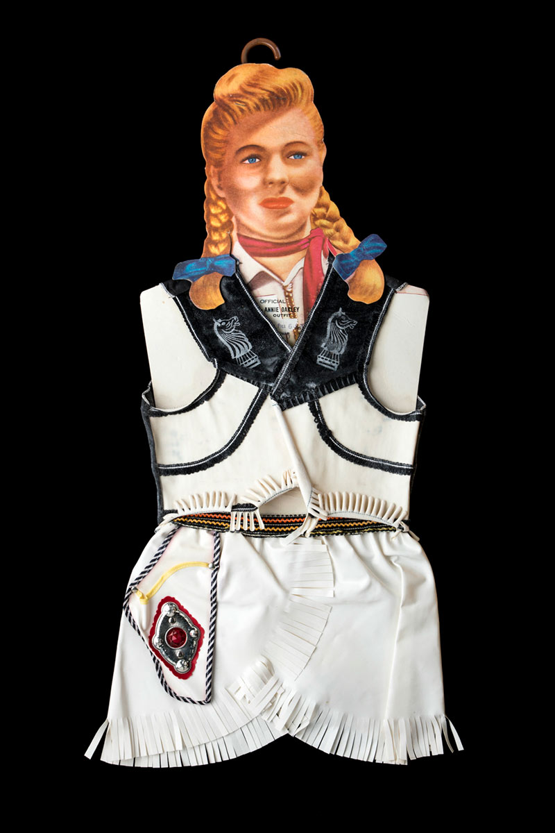 An 'OFFICIAL ANNIE OAKLEY' costume comprised of a white vest with black fringing and details, and a white skirt with a white holster trimmed in black and white fabric. The outfit is attached to a cardboard label featuring the image of a woman in pigtails, and a brown plastic hanger. The material for the vest is stiff and inflexible. - click to view larger image