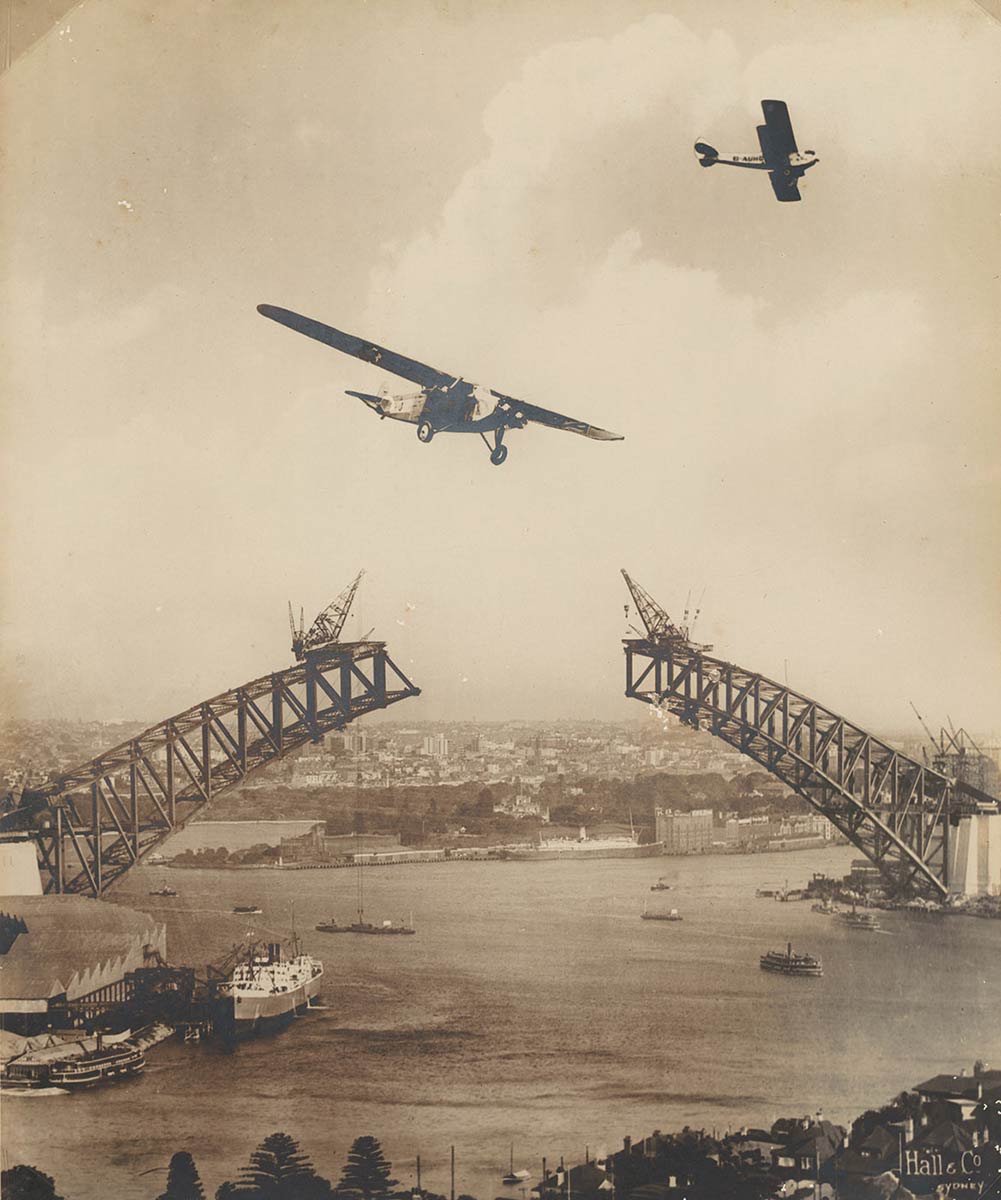 Black and white photograph showing two aeroplanes flying above Sydney Harbour and the the unjoined spans of Sydney Harbour Bridge. - click to view larger image