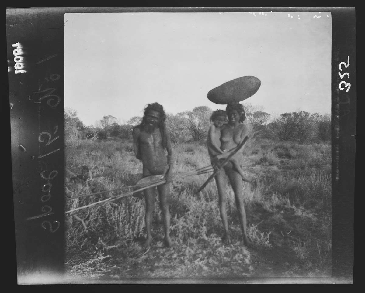 First Nations man Dinjimanne, his wife and daughter, Giles West Camp, South Australia, 1903. Dinjimanne stands to the left, holding horizontally two spears and a spear thrower in his left hand. His wife stands to the right, with their daughter on her right hip and a large wooden bowl balanced on her head. She holds what appears to be a digging stick in her right hand. Behind them is a patch of knee-high bushes leading to trees in the distance. - click to view larger image