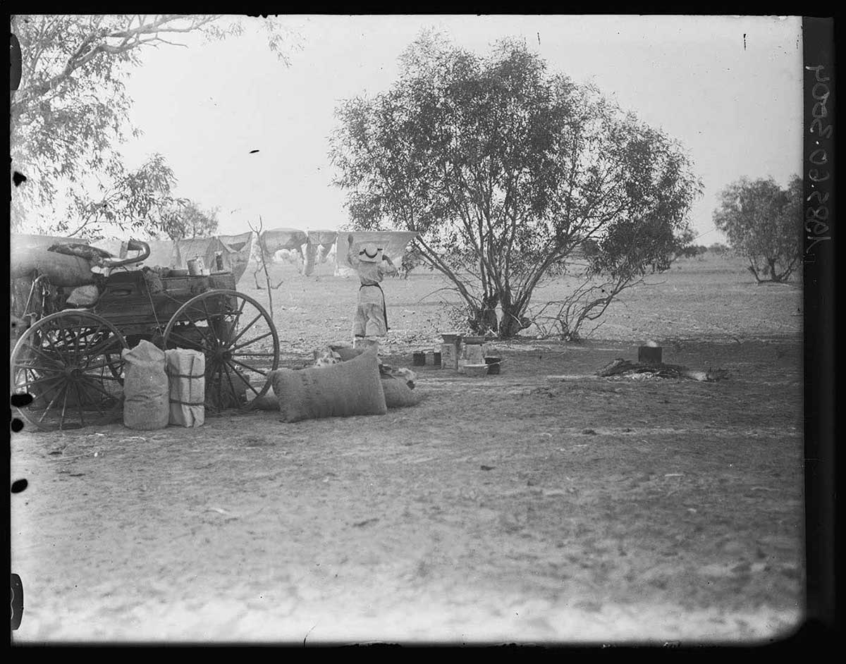 Nell Basedow hanging out the washing on a line strung between two trees, Dickerie Water Hole, South Australia 1919. She stands in the middle distance with her back to the camera. One end of the line is tied to a small tree just to her right. In the left foreground are some sacks and camping equipment on the ground next to a horse buggy. A billy boils on a fire in the right middle distance, not far from Nell Basedow. - click to view larger image
