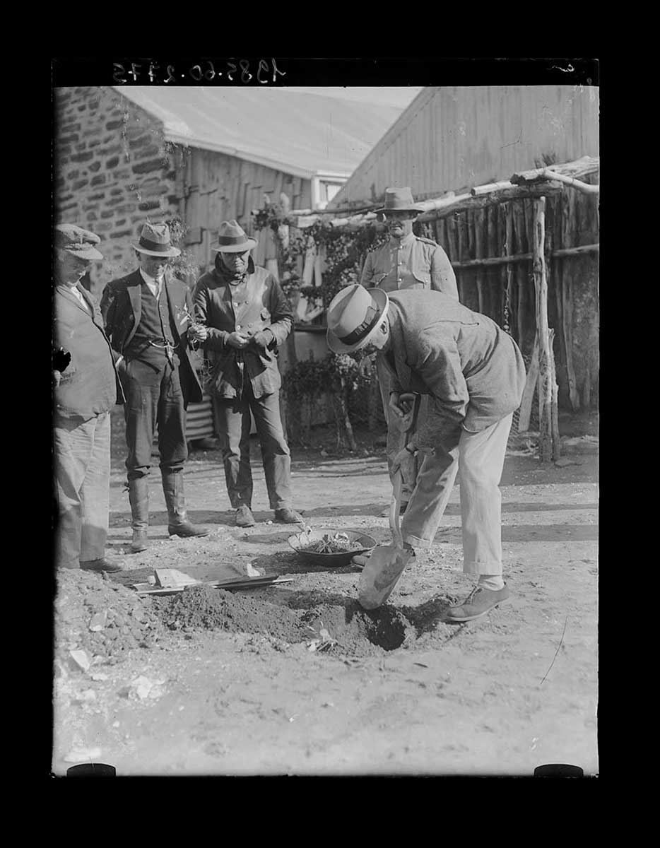 Governor of South Australia Sir Tom Bridges planting a tree, Arltunga, Northern Territory 1923. The Governor is in the right foreground, bent over digging a hole with a short-handled shovel. He is dressed relatively informally i.e. trousers, jacket and hat, with no ceremonial garb. Behind him stands a non-Aboriginal man in a uniform. His lower body is obscured by the Governor. To the left of the image stand three other non-Aboriginal men, watching the Governor. One wears a hat, leather coat and trousers, one wears knee-length boots, trousers, waistcoat, jacket and hat and the last one wears trousers, jacket and a soft peaked cap. This man stands closest to the Governor. In the background is a rough wooden fence and part of a building made from timber and bricks. - click to view larger image