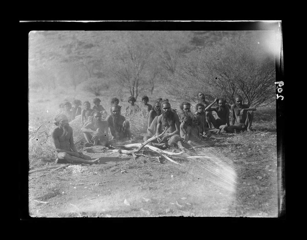 Aboriginal camp of about 20 people, McNicol Range, Northern Territory 1926. About twenty two Aboriginal peoples sit on the ground, roughly arranged in two groups. The group closest to the camera is mostly men; two children sit with them. Behind the men sits a group of women, partially obscured from the chest down by patches of scrub grass. Some of the men in the group sit around several branches laying on the ground. The ground in front of the men is thinly covered with patchy grass. Behind the women is some open scrub with scattered low bushes and trees. The image has white marks on it indicating possible damage to the print or the negative. - click to view larger image