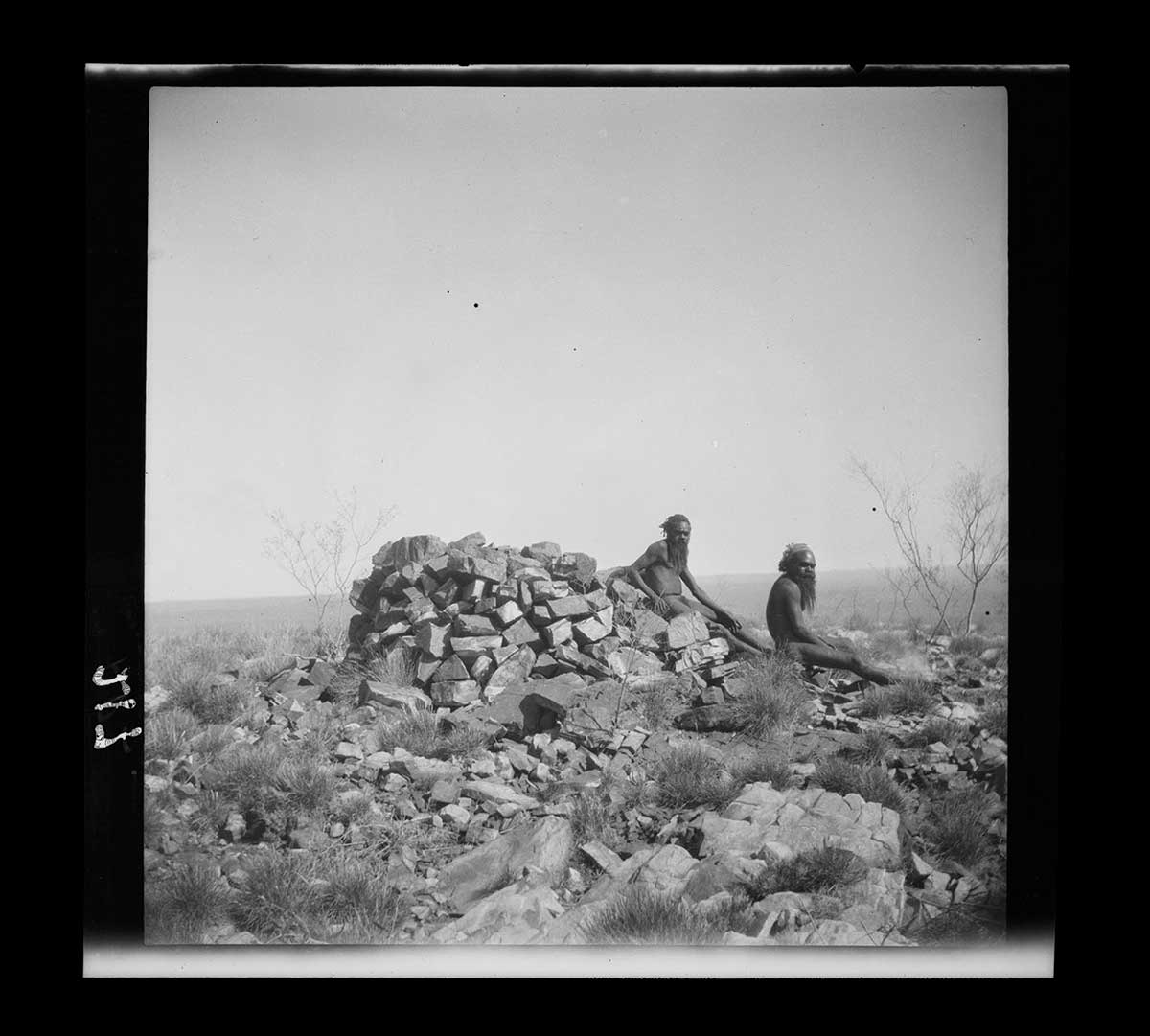 Two Aboriginal men, Artuatama and Mingerinnya, on the summit of Mount McCulloch (Nangorru), Petermann Ranges, Northern Territory 1926. The men sit to the right of a cairn of rocks in the centre of the image. One sits directly on the cairn, the other sits at its edge. Both face to the right of the image. The ground around them is covered in rocks and tufts of grass. The horizon is visible in the background. It slopes slightly down from right to left, suggesting that the camera was not level when the photograph was taken. The sky in the image appears to be compeletly cloud-free. - click to view larger image
