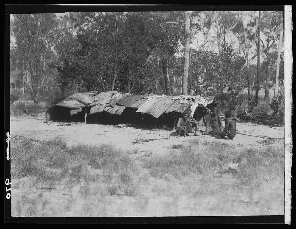 Expedition guides next to wet-weather hut, near the Liverpool River, Northern Territory 1928. Four Aboriginal adults and one Aboriginal child are at one end of a long, low rectangular floor plan hut. the adults sit in a rough circle while the child stands at the right of the adult on the right. The hut appears to be no more than about chest-high. It's covered with sheets of bark and blankets. The covering overlaps in a way designed to keep rain out. The hut has one wall and one enclosed end; the end near to the people and the long side nearby are open. The ground around the hut is bare; beyond it in the foreground are scattered grass tufts. Behind the hut is scrub with trees and taller scrub grass. - click to view larger image