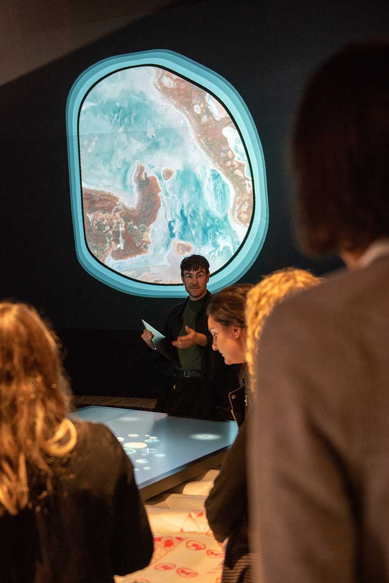 A group of people stand around a digital screen with a projection of an object on the wall behind. - click to view larger image