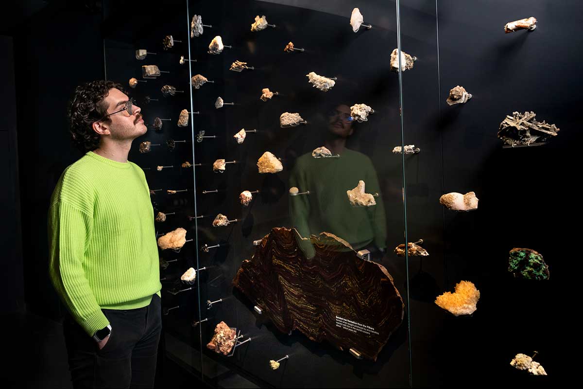 A man wearing a lime green jumper stands with his hands in his trouser pockets, gazing at a museum display case filled with mounted mineral and rock specimens of various colours and textures. - click to view larger image