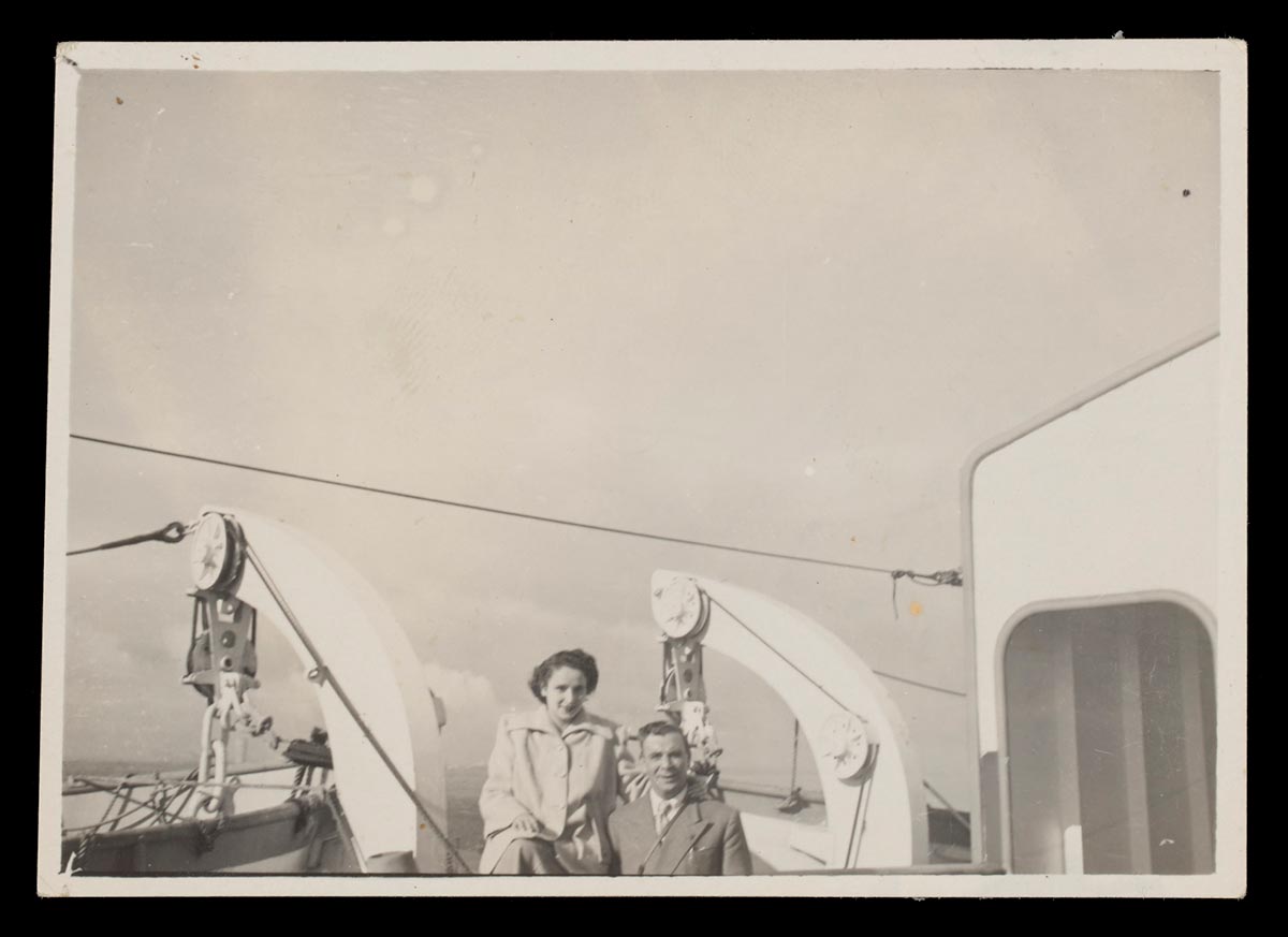 Landscape-oriented black and white photograph of Effie and Peter Kyprios on the deck of a passenger ship. The couple are positioned at the bottom of the photo, with lifeboat rigging behind them and open sky above them. They are both wearing coats and smiling towards the camera. - click to view larger image
