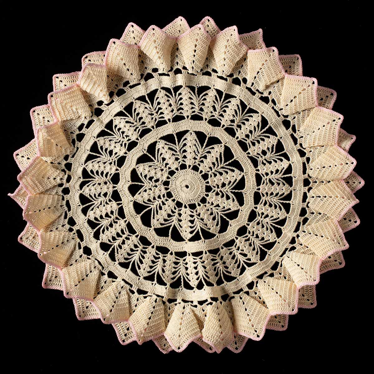 Circular off-white crocheted doily with three-dimensional edges creating a frill-like effect on a black background. The frills are edged in light pink thread. The centre of the doily is circular, with triangular crocheted designs in concentric circles creating a sunflower effect. - click to view larger image