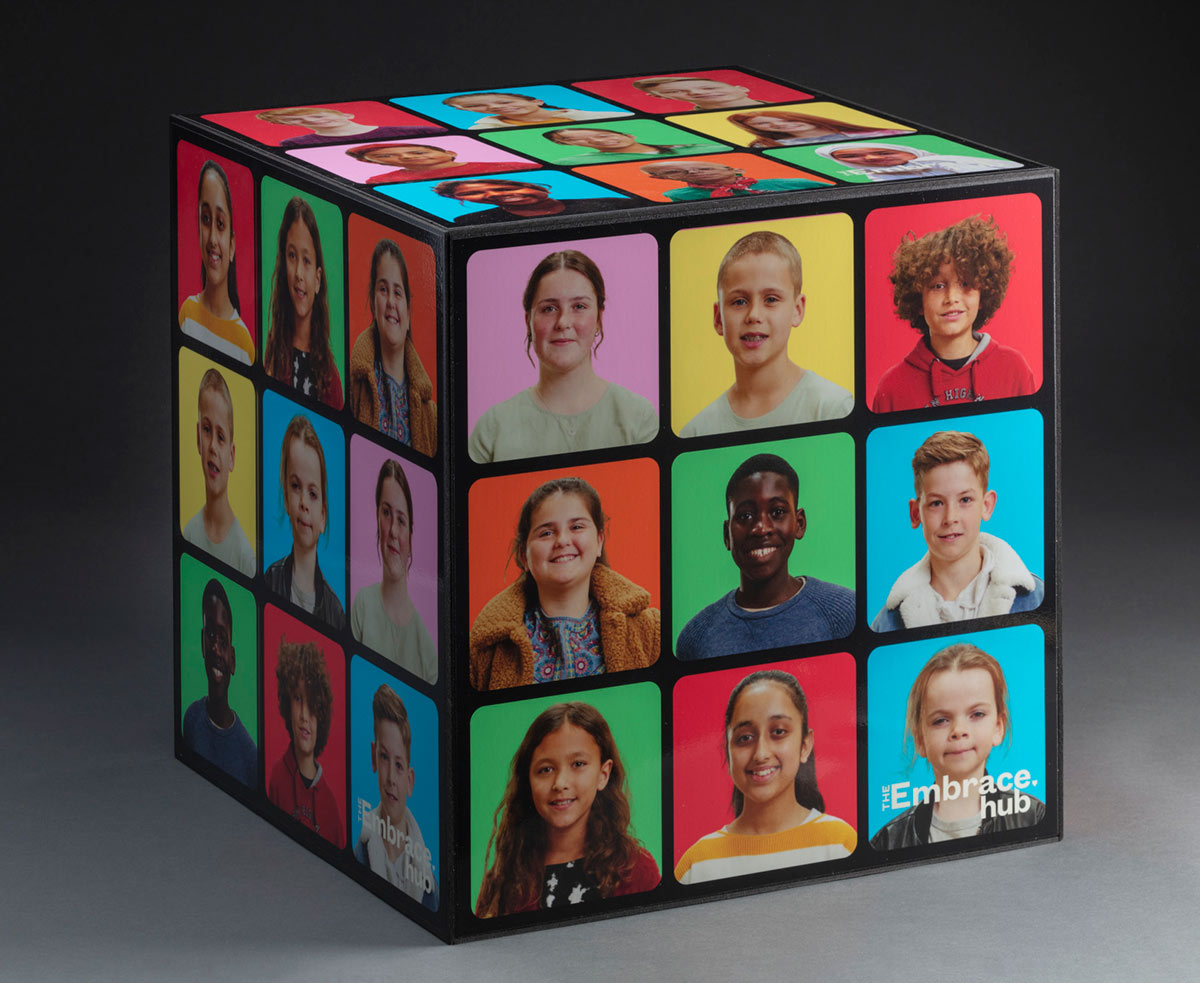 An artwork in the form of a cube with a set of nine portraits on each side accompanied by the words 'THE / Embrace / hub'. Each portrait has a plain background in one of six colours, including blue, green, red, orange, yellow and pink.
