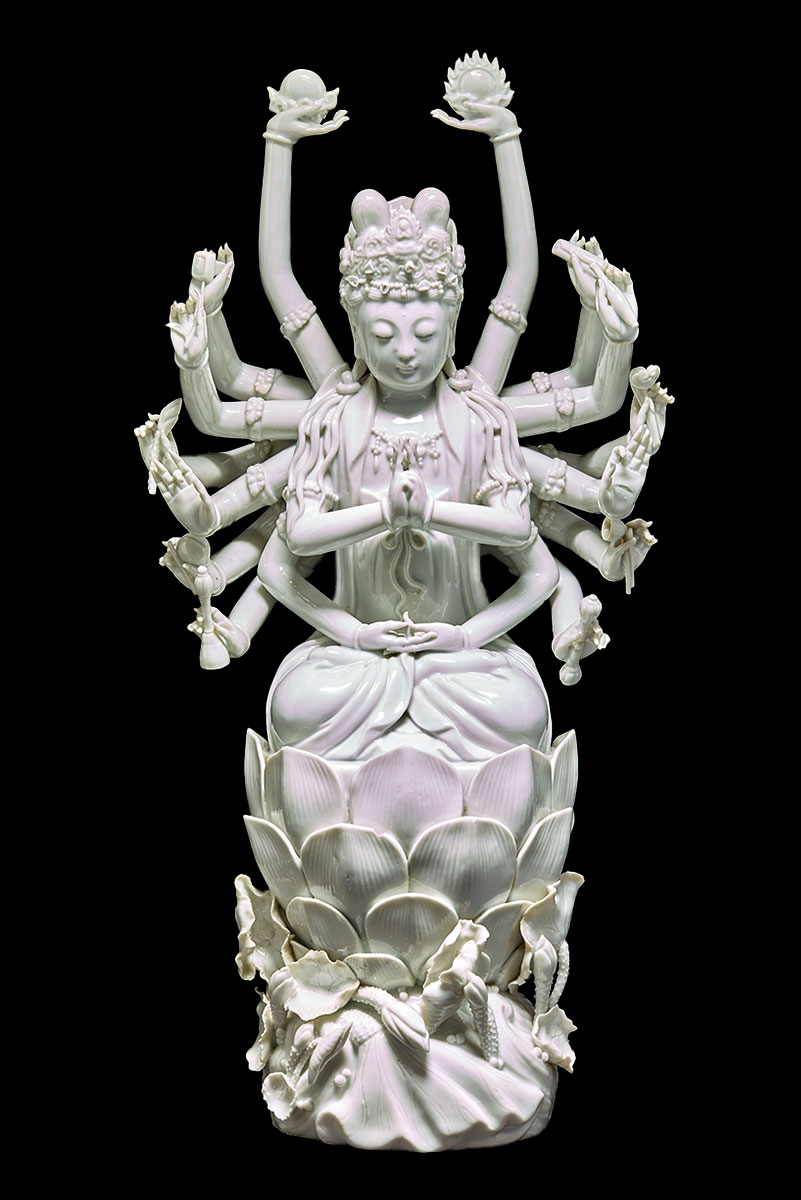 Porcelain sculpture of a figure with eighteen arms, seated on a ornately decorated base. - click to view larger image