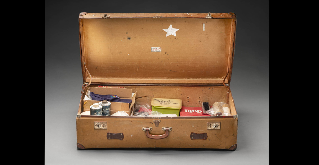 A photograph of Norman Hetherington’s suitcase which he took to the studio when recording. The suitcase is brown with a leather handle and two lockable metal latches. The contents of the suitcase is: three books, a brush, four plastic containers and contents, an electrical cable, fishing line, a folder of riddles, gloves, two hats, a hook, painting equipment, puppet parts, five repair kits with various contents, ropes, two cardboard signs, three bandanas, a piece of fabric, two tins and contents, a toy flute, two umbrellas and various other components for the repair of puppets.