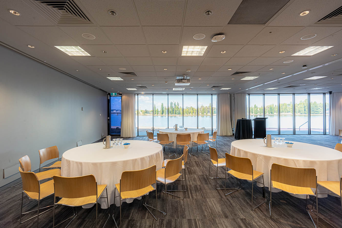 A photograph of the Museum's Peninsula room. The room has a large window at the back which looks out to the lake. The room has large tables with white table clothes and chairs set up for a function. 