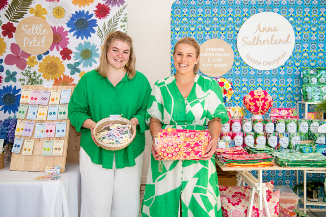 Two women stand displaying a basket of colourful earrings and small textile bags.. - click to view larger image