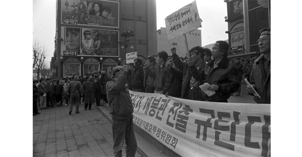 A reproduced black and white image of filmmakers protesting the direct distribution of foreign films, outside and in a public space on 1 December 1990. A large group of individuals stand at the front holding a long protest banner. with Korean text They are tightly packed, and appear to be shouting slogans. One man faces the crowd, recording the protest. In the background, tall buildings are adorned with large movie posters.