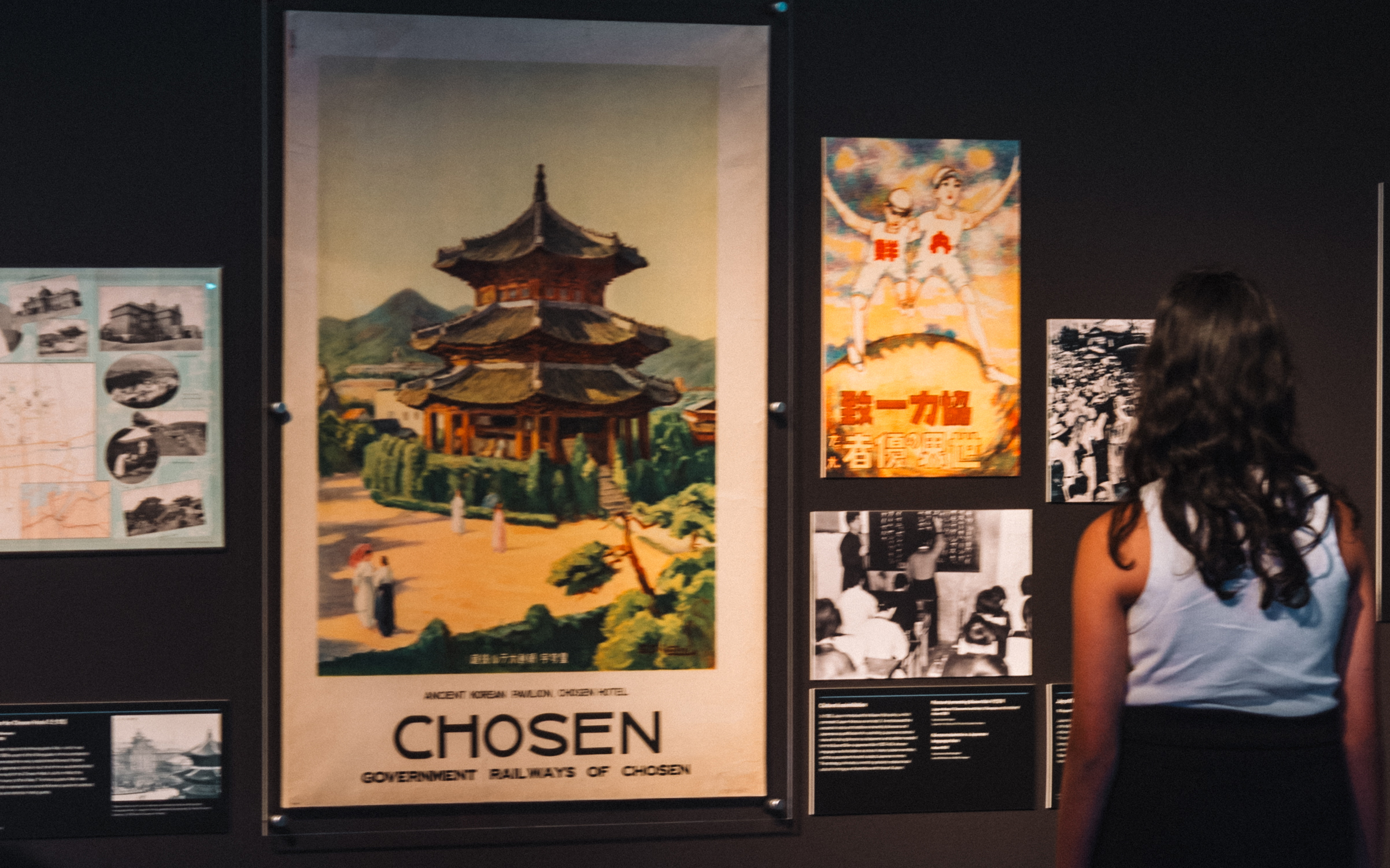 A photograph of a young person wearing a white top and black skirt. They are looking at a historic Korean poster.