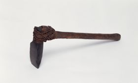 Small stone axe with a handle made from medium brown hardwood and a blade made of fine-grained black basalt both tied together with coconut fibre. The end of the T-shaft appears to be broken off.