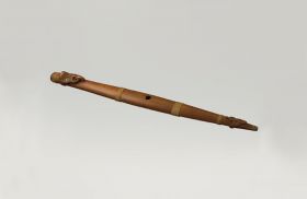 Flute made of wood with two halves mounted together. A carving at the lower closed end and the upper open end is a human head with mother-of-pearl eyes and an extended tongue.