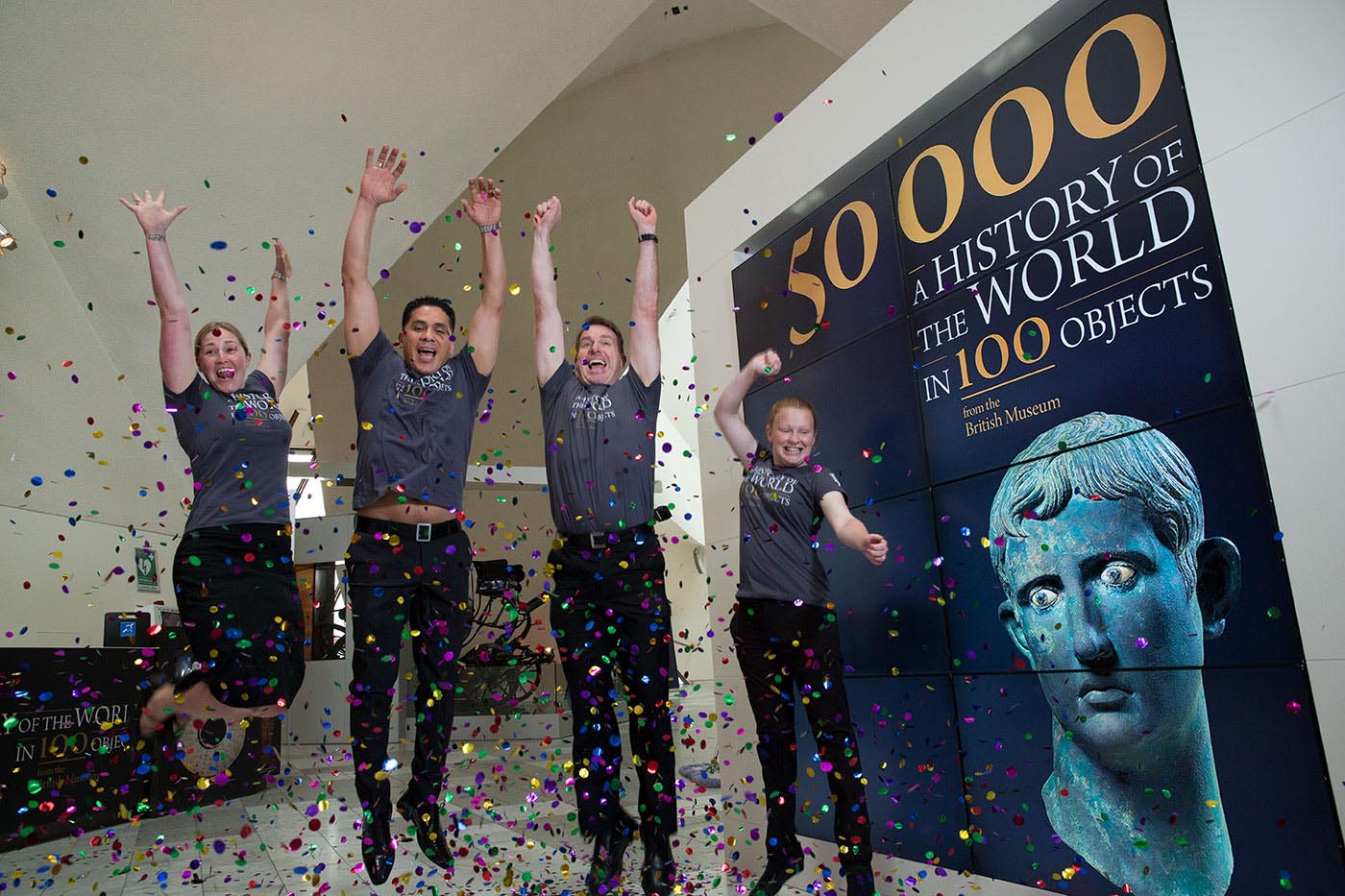 Two women and two men in uniform jumping in the air amidst confetti, adjacent to a promotional billboard.