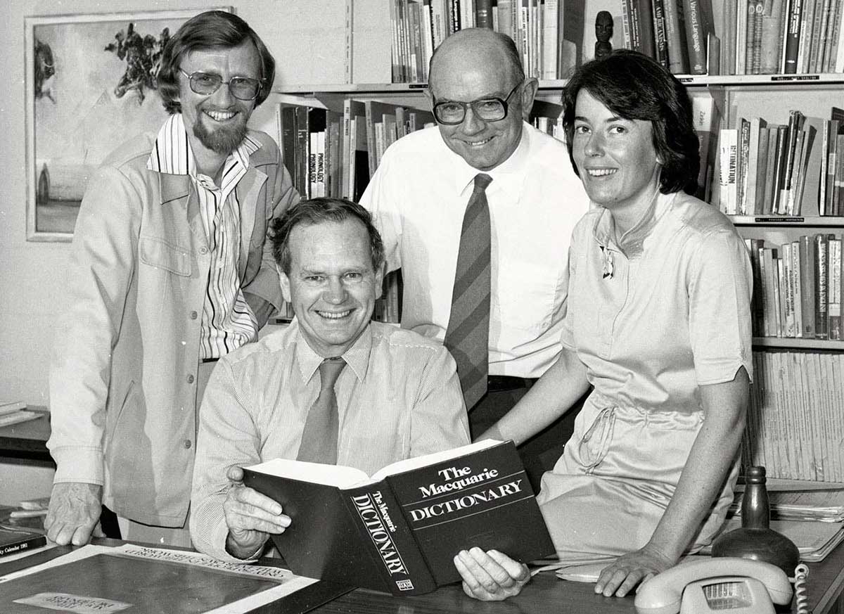 A black and white photo of four people inside an office. There is a bookcase behind them and the man seated is holding a Macquarie Dictionary.