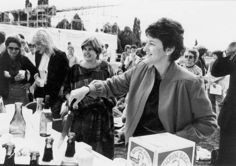 Black and white photograph of a woman smiling at outside event. Several women are in the background. Senator Ryan is holding a knife for cutting a cake. Champagne bottles are visible. - click to view larger image