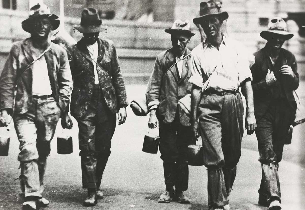 Black and white photograph of five men walking towards the camera dressed in shabby clothes. Most are carrying swags and billy cans.