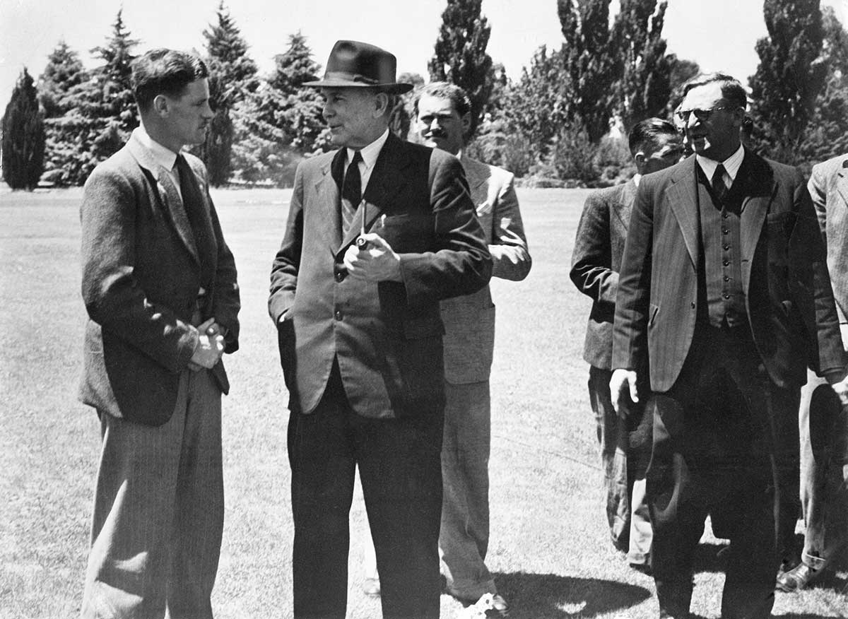 Outside photo of young man with folded hands talking to a smiling Prime Minister Chifley. Calwell stands to one side.