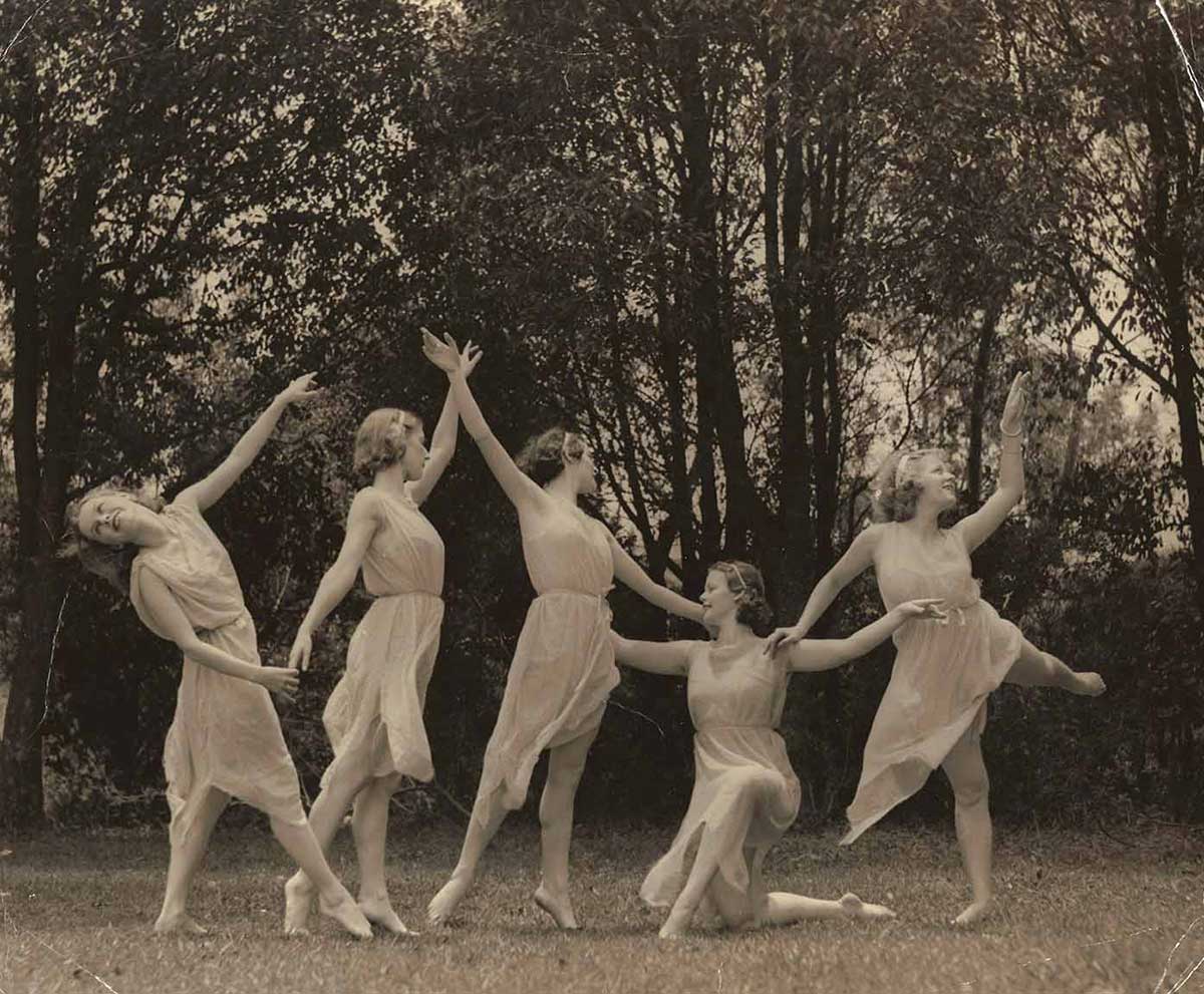 Five ballerinas dressed in white dresses perform on what appears to be grass with trees in the background.