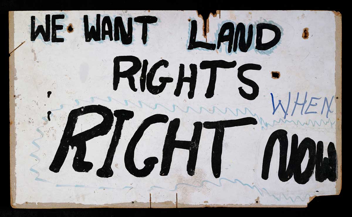White sign with the words ‘We want land rights. When. Right now’ hand painted in black.