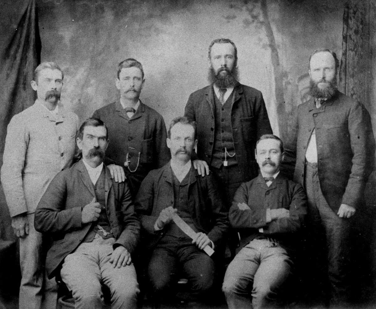 Seven men in a studio group photograph, staring unsmiling at the camera. The name of organisation has been hand-written on the top frame.