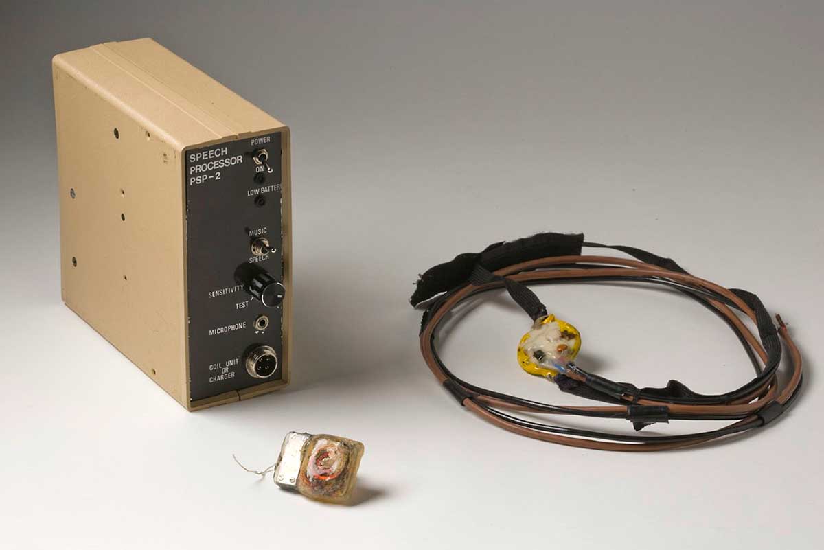 Small bionic ear implant, with coiled cables inside, pictured with a box marked 'Speech Processor PSP-2' and a small receiver unit attached to coiled cables and a velco headstrap.