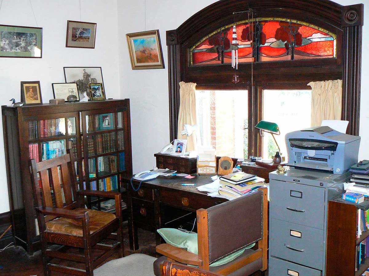 A room with a table, bookcase, filing cabinet and two chairs. The table faces a window that has a red, blue and purple stained glass feature above it. On the table are books, boxes, a tall lamp, a modern keypad telephone and loose papers. There are photographs on the bookcase and paintings hung on one wall.