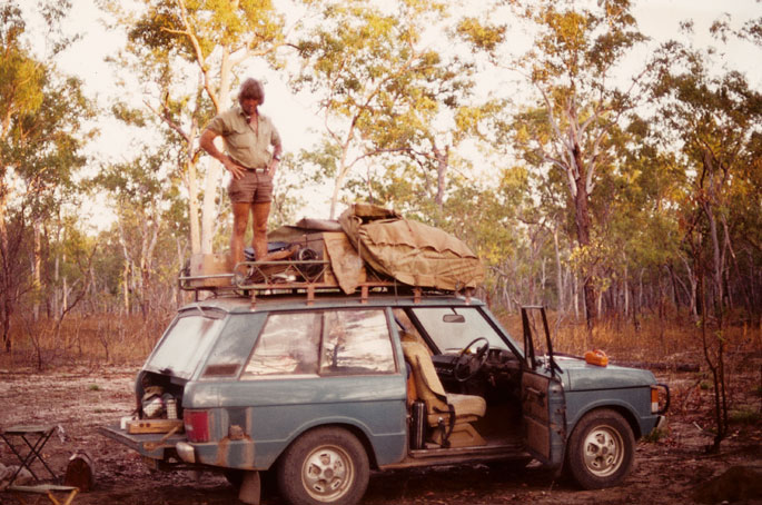 Fred Hollows, in khaki shirt and shorts, stands on the roofracks of a four-wheel-drive vehicle. Various equipment is packed onto the roofracks. The vehicle is parked in a clearing surrounded by tall trees. - click to view larger image