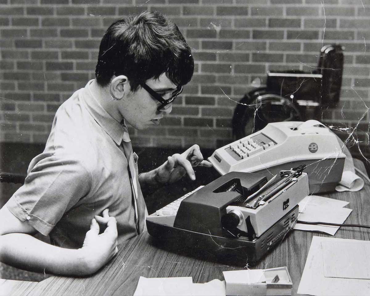 Black and white photograph showing a young man seated at a table, typing with his left hand. - click to view larger image