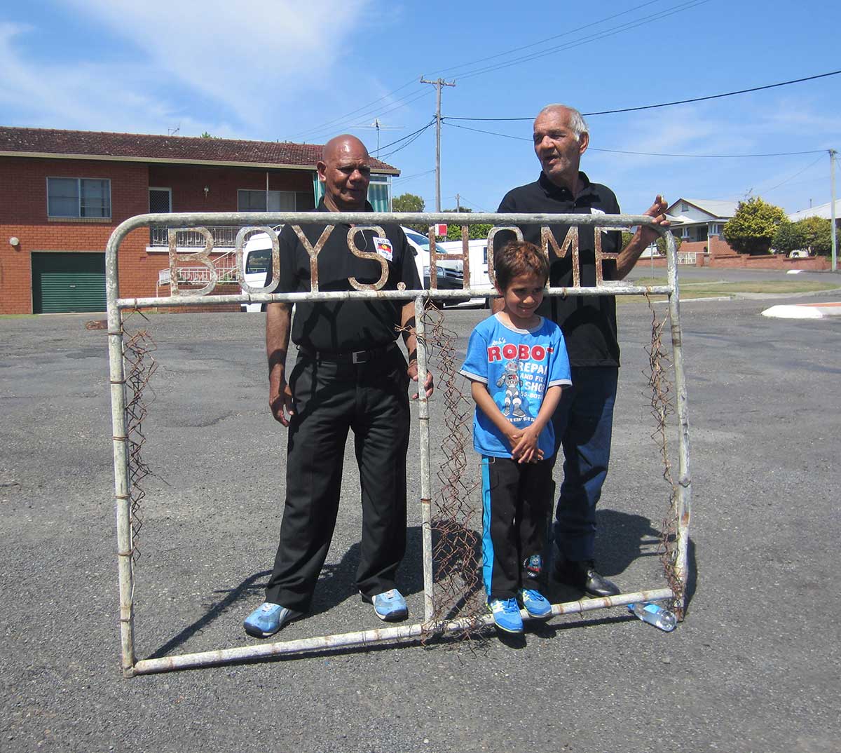 Colour photograph of two men and a child posing with an old metal fence with signage that reads: 