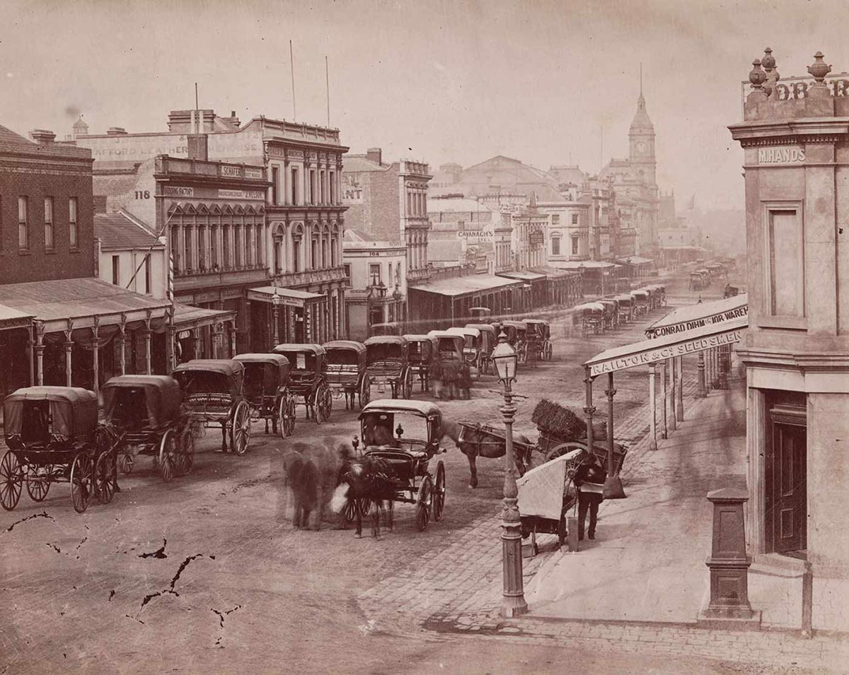 Black and white image of an urban streetscape with a series of two-storey buildings on either side. Many have verandahs at the front and a buildig with a spire rises towards the far end of the street. A series of horse-drawn carriages is lined up along the left side of the street. Another two carriages and several people stand to the right.