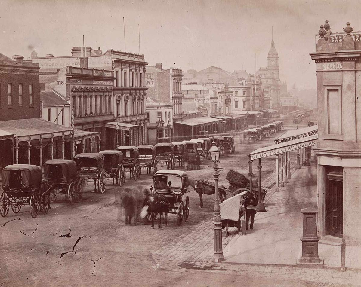 Black and white image of an urban streetscape with a series of two-storey buildings on either side. Many have verandahs at the front and a buildig with a spire rises towards the far end of the street. A series of horse-drawn carriages is lined up along the left side of the street. Another two carriages and several people stand to the right. - click to view larger image