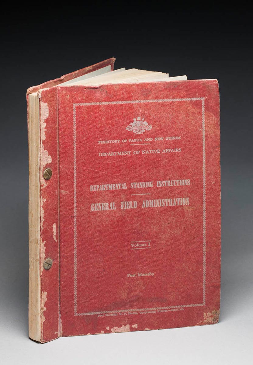 A red hardcover book with a loose leaf inside the front cover. The book features two gold coloured fixings through spine, and silver coloured text on the front cover, which is within a silver border and reads 'TERRITORY OF PAPUA AND NEW GUINEA / DEPARTMENT OF NATIVE AFFAIRS / DEPARTMENTAL STANDING INSTRUCTIONS / GENERAL FIELD ADMINISTRATION / Volume 1 / Port Morseby'. - click to view larger image